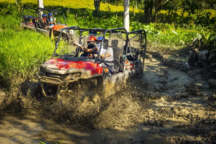 ATVs driving through muddy terrain with splashing mud in a grassy area with trees.
