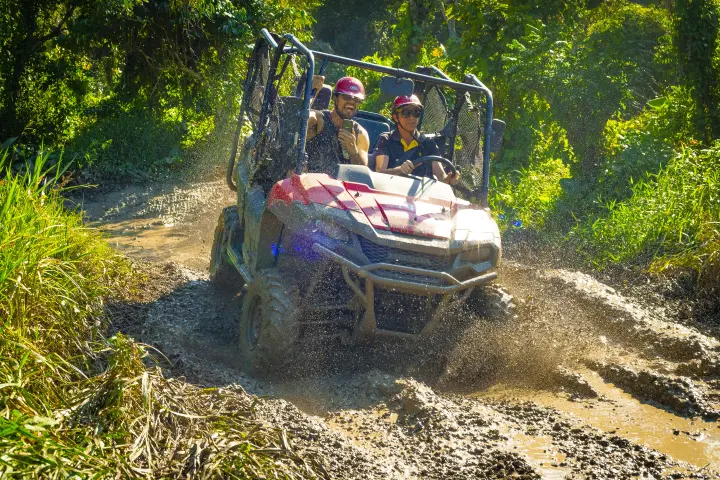 Two people driving a red ATV through a muddy trail surrounded by greenery.
