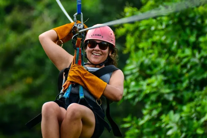 Person ziplining through lush green forest, wearing helmet and gloves.