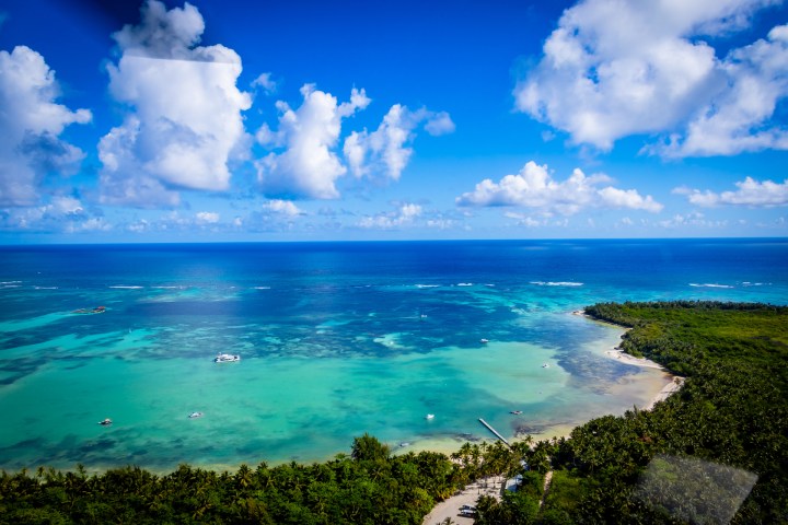 Aerial view of turquoise ocean, sandy coastline, and green vegetation under a blue sky with clouds.