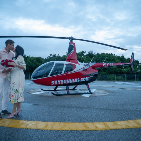 Couple kissing near a red helicopter on a helipad.
