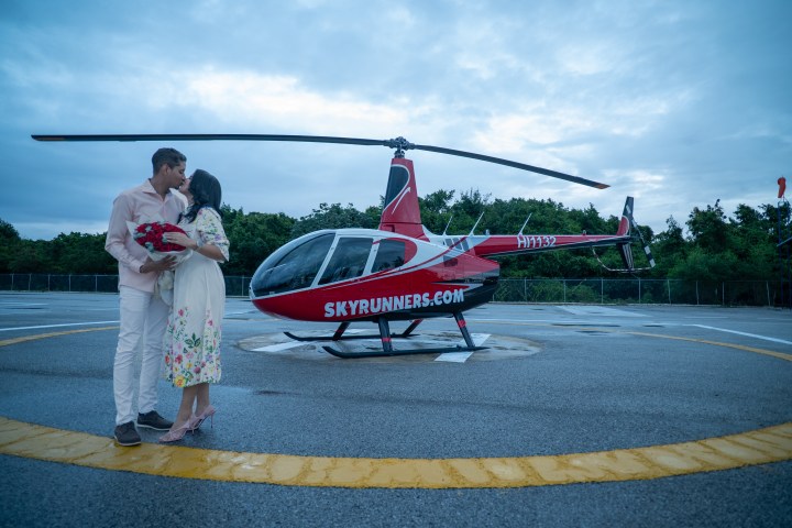 Couple kissing near a red helicopter on a helipad.