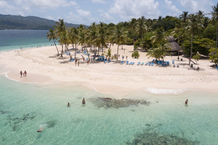 Beach with palm trees, loungers, and swimmers in clear water under a bright sky.