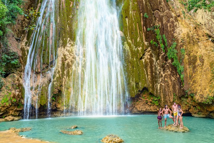 Four people standing near a waterfall cascading into a turquoise pool surrounded by rocky cliffs.