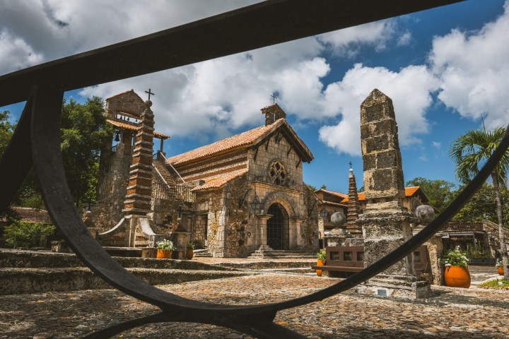 Historic stone church with bell tower, framed by wrought iron, under a blue sky with clouds.
