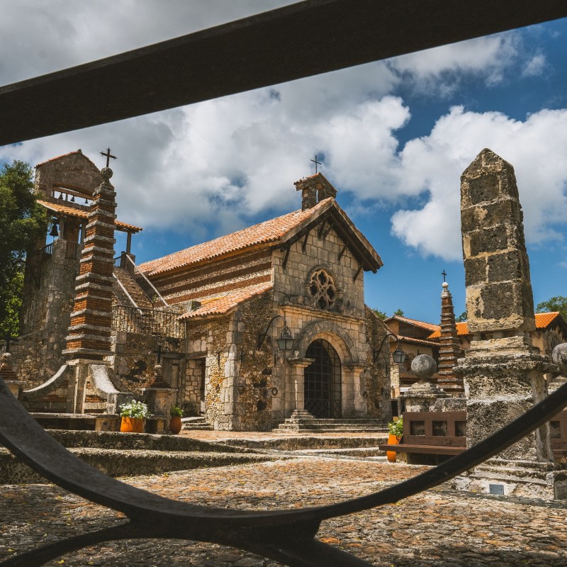Historic stone church with bell tower, framed by wrought iron, under a blue sky with clouds.