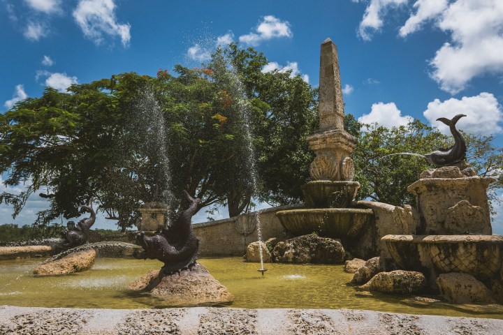 Stone fountain with dolphin sculptures spraying water, trees and blue sky in the background.