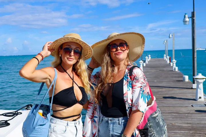 Two women in sun hats and sunglasses standing on a pier with the ocean in the background.