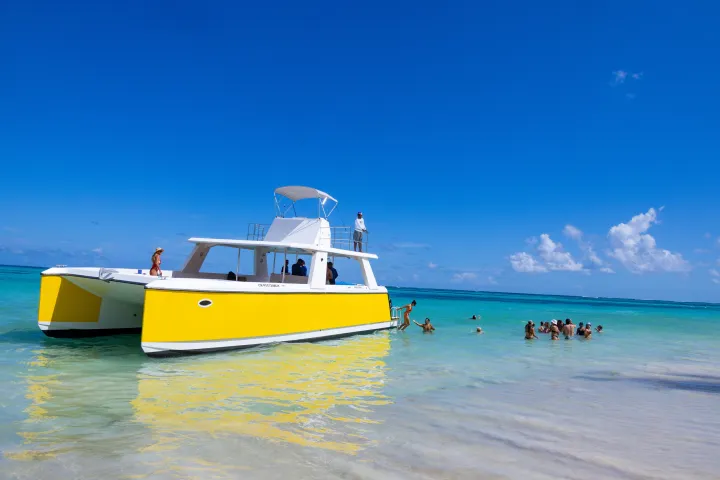 Yellow catamaran near the shore with people swimming in clear blue water under a clear blue sky.