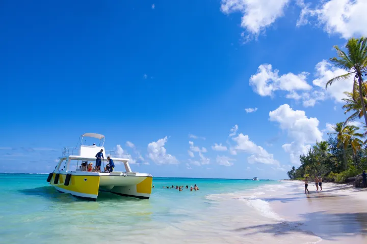 Catamaran near a tropical beach with palm trees and people swimming under a clear blue sky.