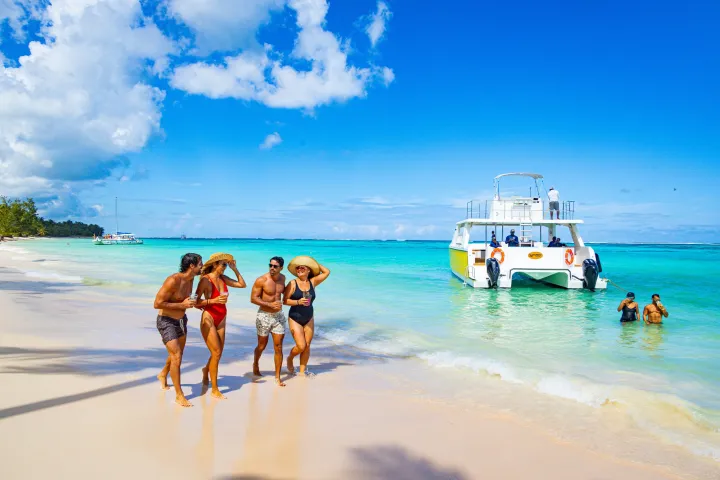 Four people walking on a beach near a catamaran in turquoise water under a clear blue sky.