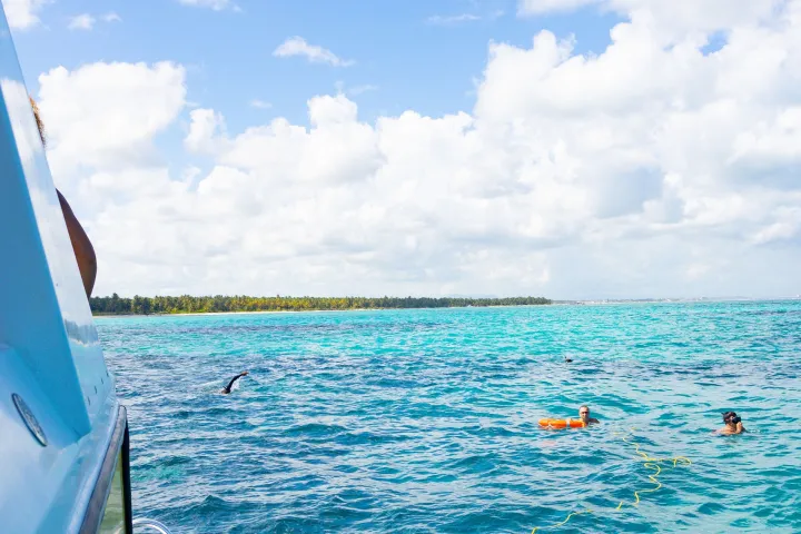 Person climbing boat ladder in blue ocean, with others swimming nearby under a clear sky.