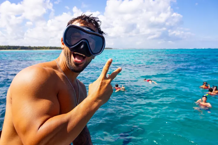 Man with snorkel gear making peace sign on boat, people snorkeling in turquoise sea.