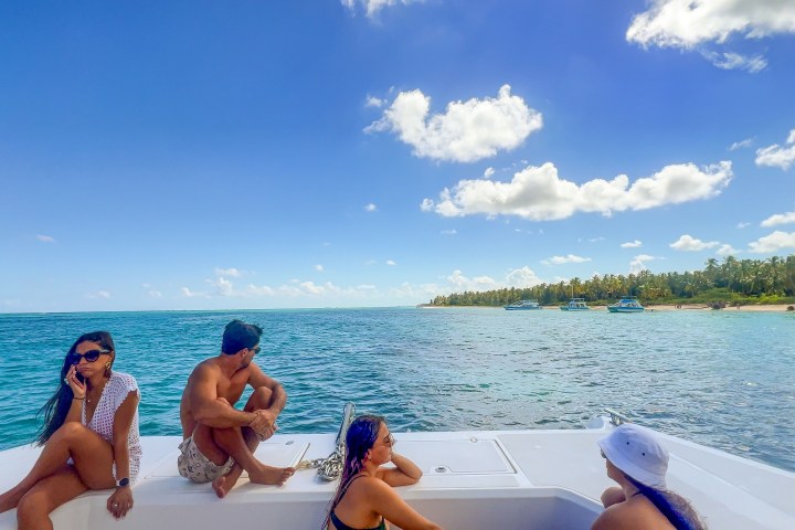 People relaxing on a boat near a tropical shoreline with clear blue water and sky.