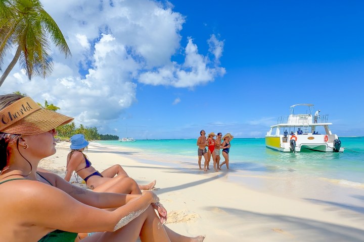 People relaxing on a tropical beach with a boat anchored nearby under a clear blue sky.