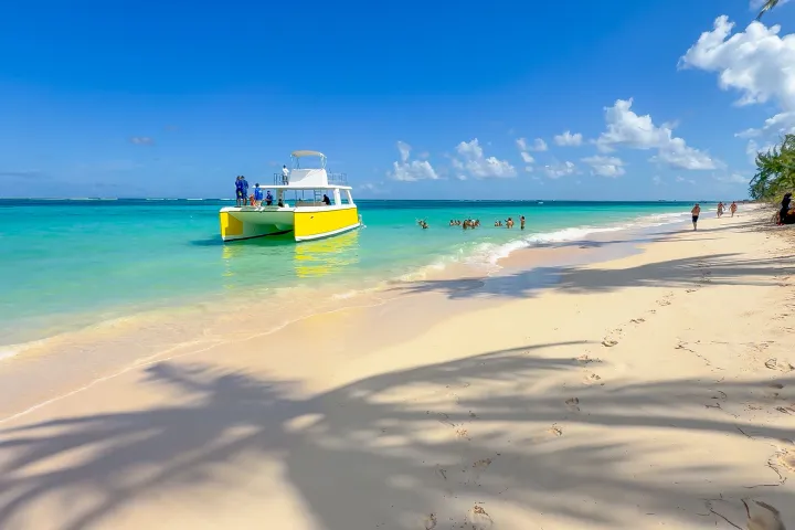 Tropical beach with yellow boat, turquoise water, people swimming, and palm tree shadows on sand.