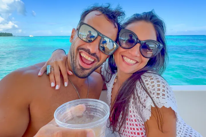 Smiling couple wearing sunglasses on a boat with turquoise sea and blue sky in the background.