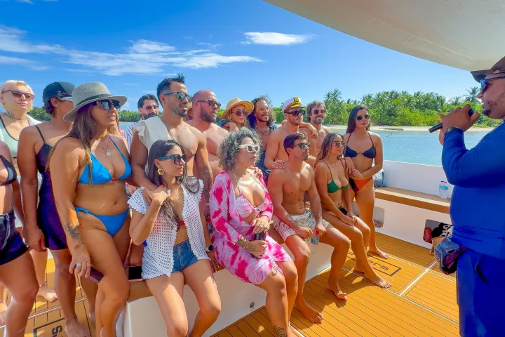 A group of people in swimwear on a boat, listening to a person speaking into a microphone.