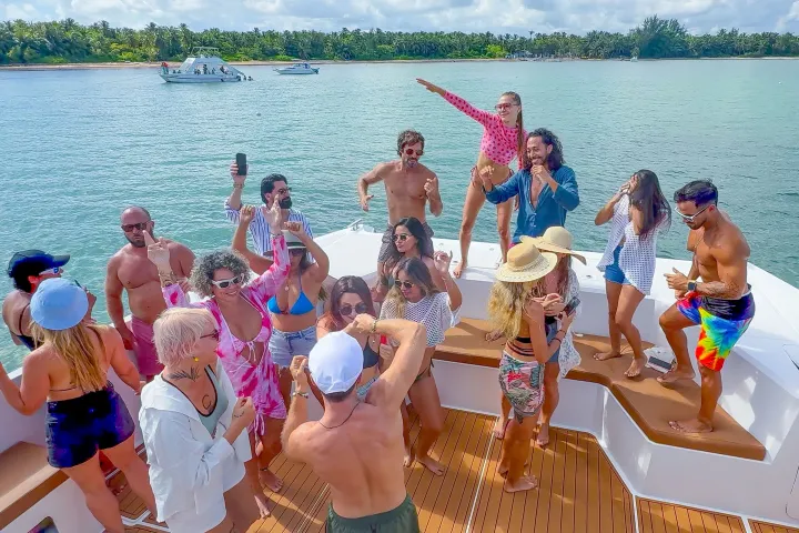 Group of people dancing on a yacht in sunny weather with palm trees in background.