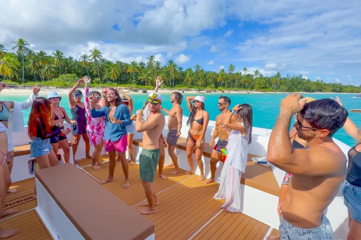 Group of people dancing on a boat near a tropical beach with palm trees and blue water.