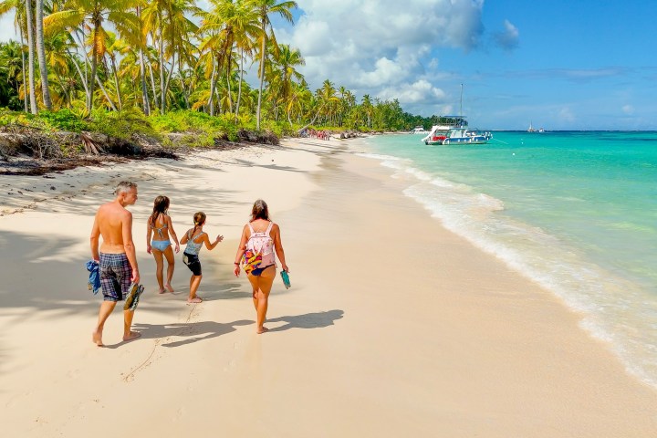 Family walking along a sandy beach with palm trees and boats on a sunny day.