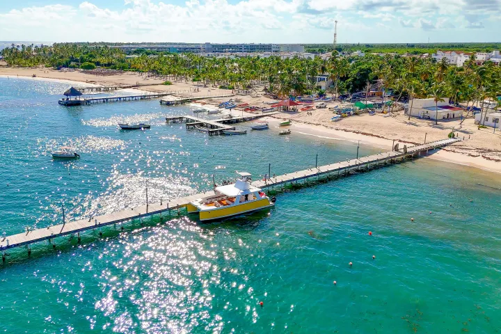 Aerial view of a beach with a long pier, boats, and palm trees under a blue sky.