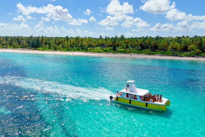 A yellow boat with people cruises on clear turquoise water near a palm-lined beach.