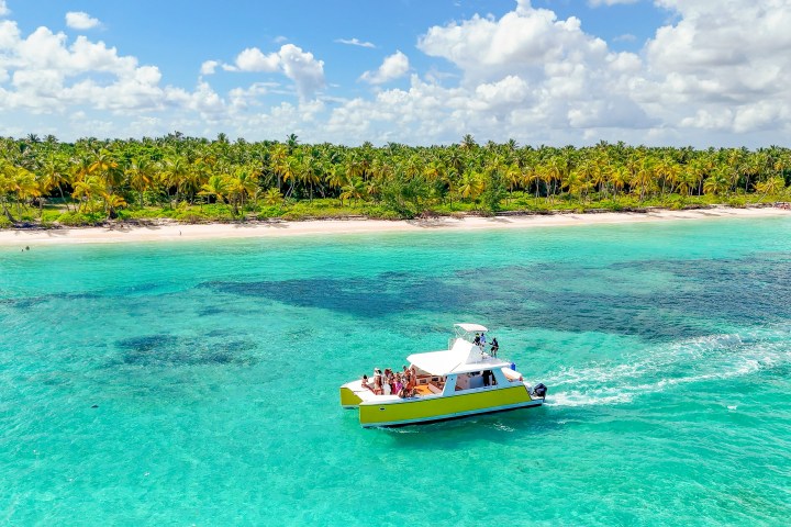 Yellow boat with people on blue water near palm-lined beach under cloudy sky.