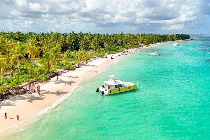Tropical beach with palm trees, turquoise water, and a boat near the shore under a cloudy sky.