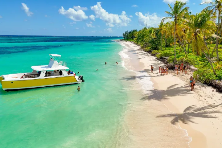Yellow boat near tropical beach with palm trees, turquoise water, and people walking on the sand.