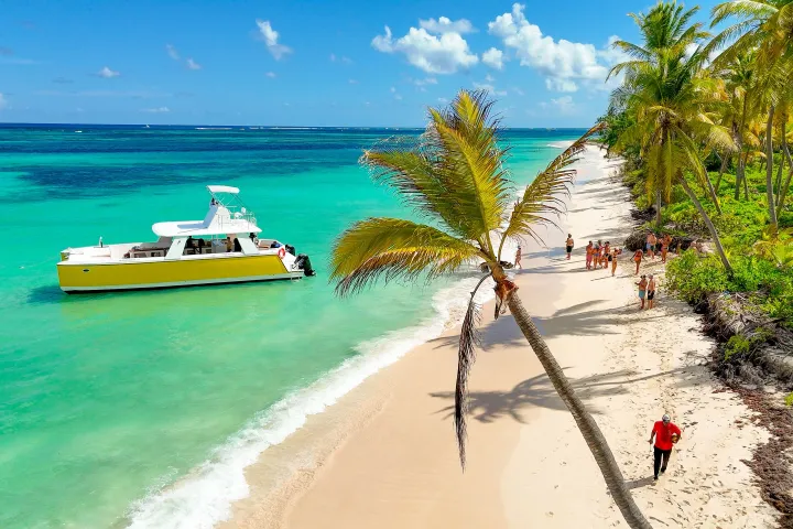 Tropical beach with people, a boat in turquoise water, and leaning palm tree.